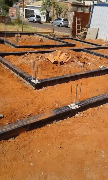 Vigas baldrame de um sobrado em obra revestidas de manta asfáltica preta, com os ferros de arranque para cima, em terreno de barro vermelho