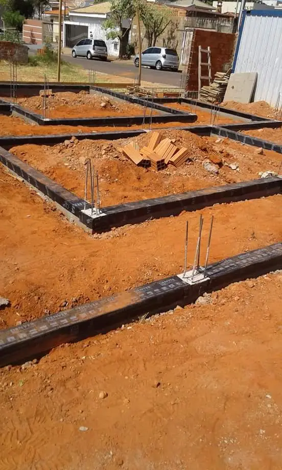 Vigas baldrame de um sobrado em obra revestidas de manta asfáltica preta, com os ferros de arranque para cima, em terreno de barro vermelho