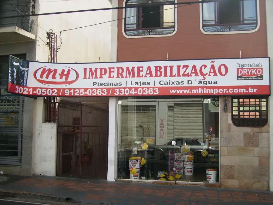 Fachada da loja MH Impermeabilização em 2009: letreiro vermelho e branco com o logotipo, “Piscinas, Lajes, Caixas d’água” e três telefones, porta de vidro com baldes de impermeabilizante na vitrine