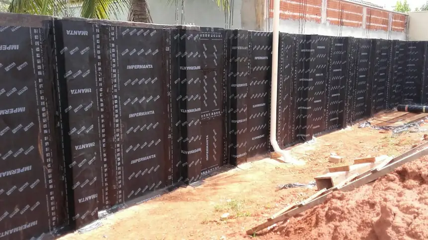 Muro de arrimo comprido inteiramente revestido de manta asfáltica preta com a estampa branca do fabricante, com o terreno ainda aberto ao pé