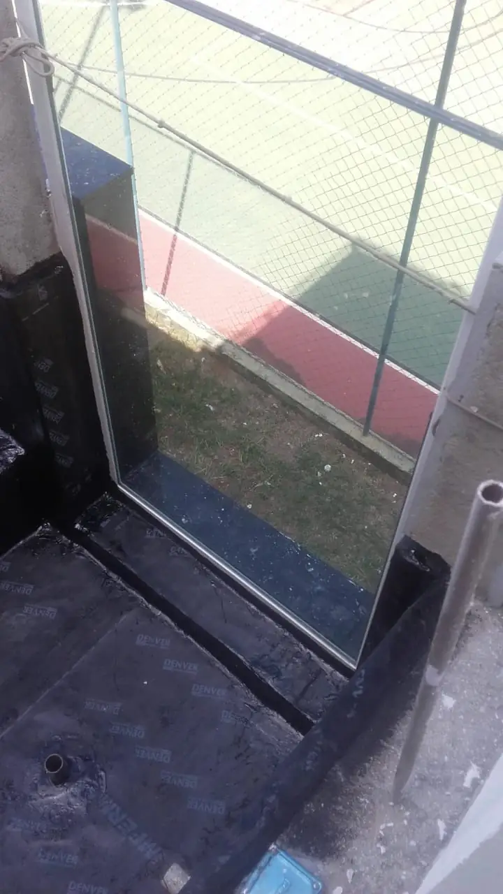 Piso com manta asfáltica subindo na base da parede junto a uma porta de vidro, com uma quadra esportiva ao fundo